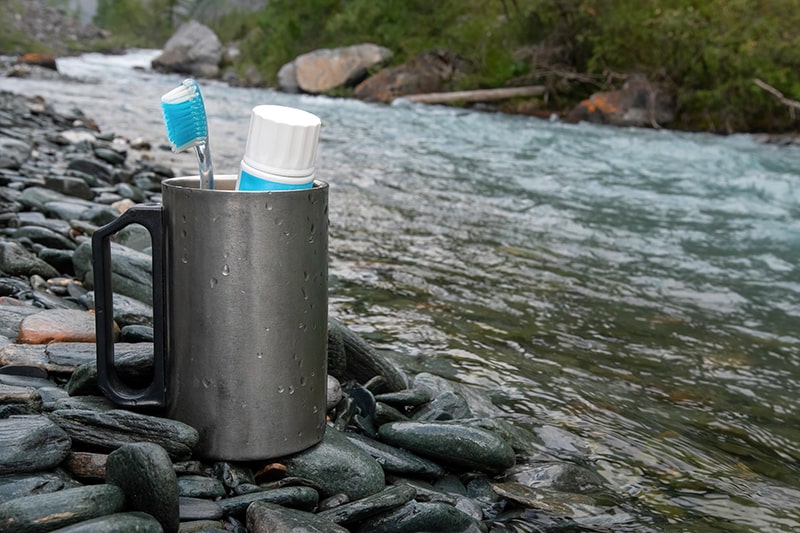 How to Stay Clean When Hiking — Camping Mug with Toothbrush and Toothpaste on the Bank of a Mountain River.